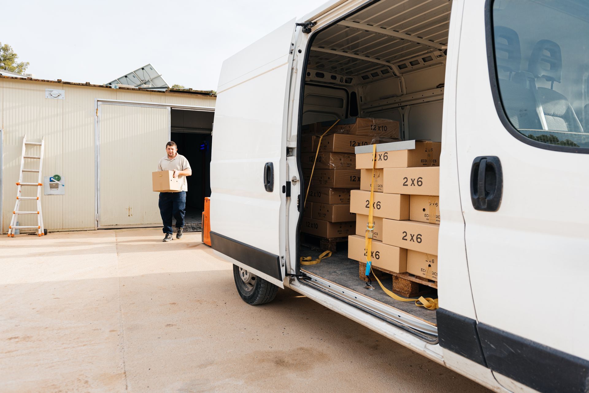 A person carries a cardboard box from a warehouse toward the open side door of a white cargo van filled with boxed goods.