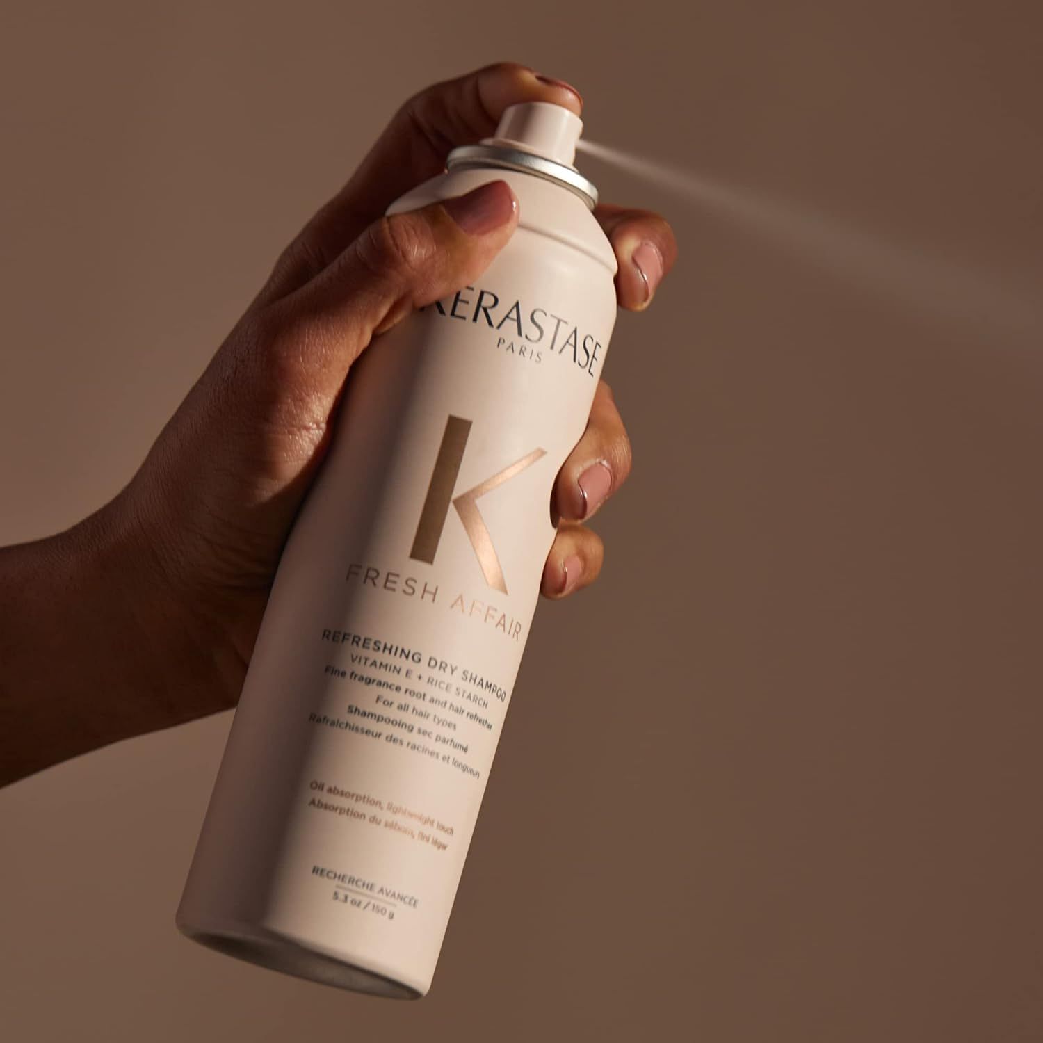 A hand spraying Kerastase hair product. The can is beige with gold lettering.