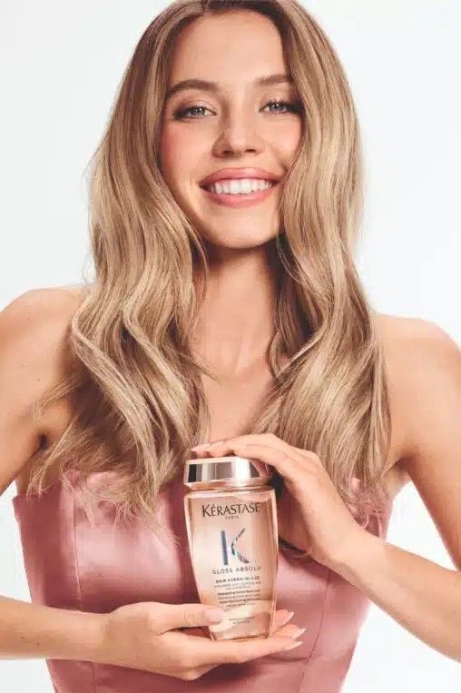 Woman smiling, holding a Kerastase shampoo bottle. Blonde hair, pink top, white background.