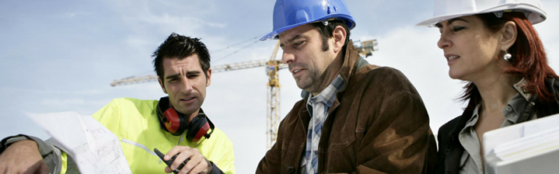 Three construction workers in hard hats examine blueprints at a building site with a crane in the background.