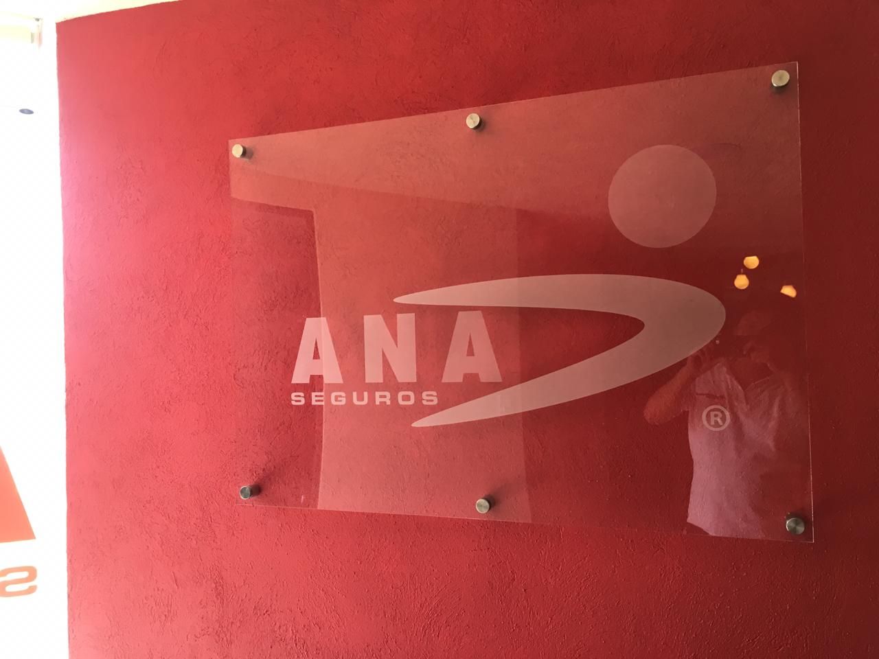 Letrero de ANA Seguros, con el logotipo esmerilado sobre un panel de vidrio, montado sobre una pared roja. Una persona se refleja en el cristal.