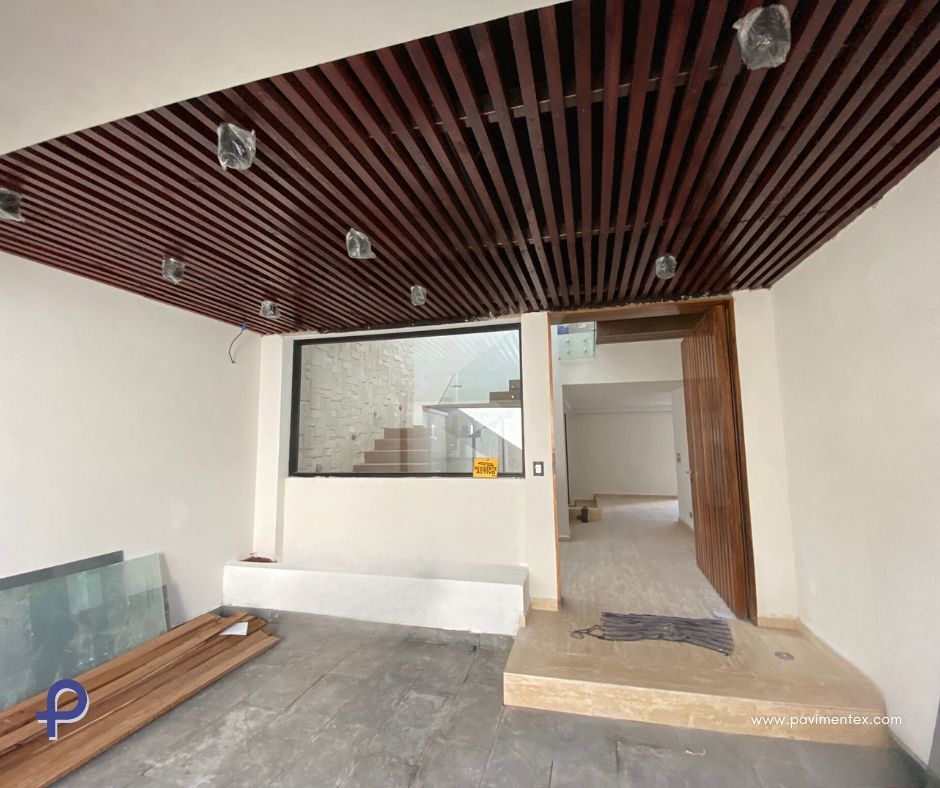 Interior de un edificio moderno en construcción; techo de madera, ventana grande, puerta, paredes blancas, suelo gris.