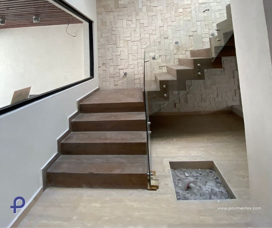 Escaleras de madera con barandilla de vidrio en una casa moderna, pared de piedra texturizada y espacio abierto debajo.