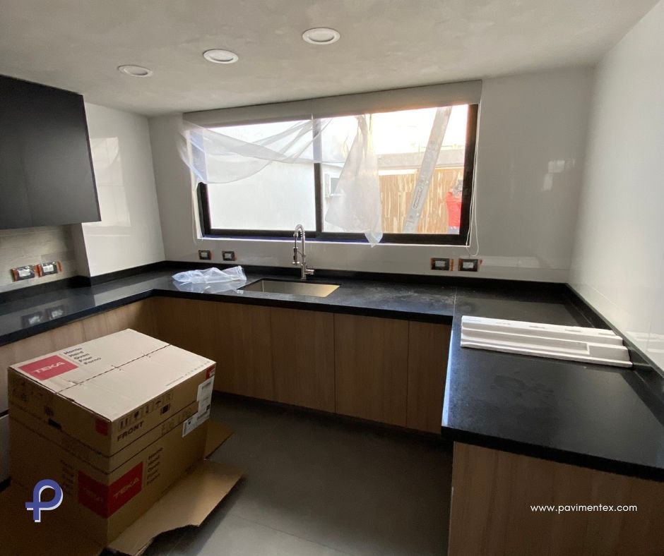 Cocina con gabinetes de madera clara, encimeras oscuras y campana extractora negra; ventana con entrada de luz.