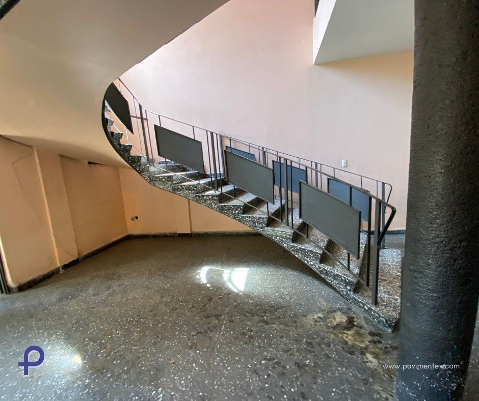 Una escalera curva con barandillas y escalones negros. Las paredes son rosas y el suelo moteado.