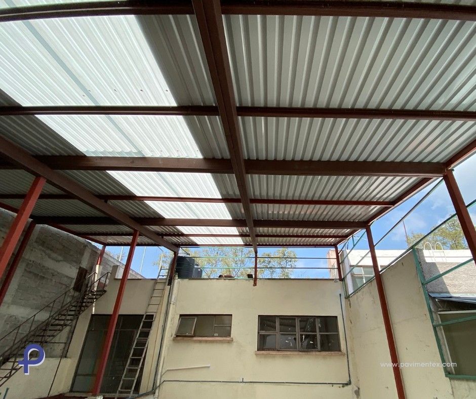 Techo de estructura de acero con chapa ondulada y paneles translúcidos. Edificio beige abajo, exterior, cielo nublado visible.