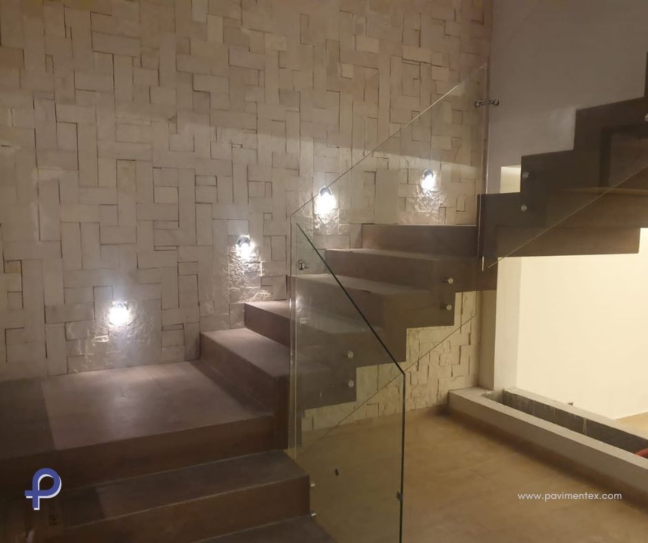 Escaleras de madera con barandilla de cristal, iluminadas por apliques de pared, junto a una pared texturizada de color beige.