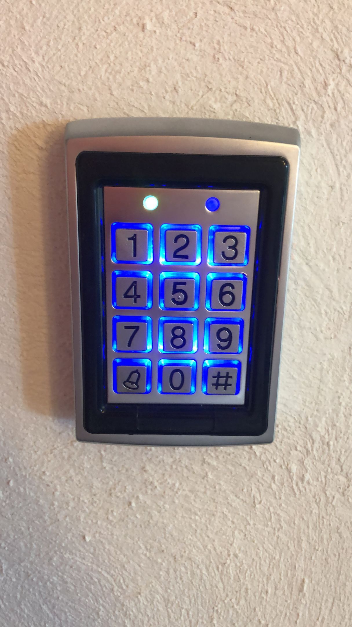 Teclado con botones retroiluminados en azul, marco plateado, montado en pared blanca.