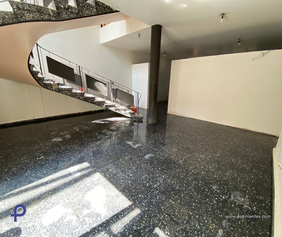 Interior con suelo moteado oscuro, escalera, columna y paredes blancas. Luz natural.