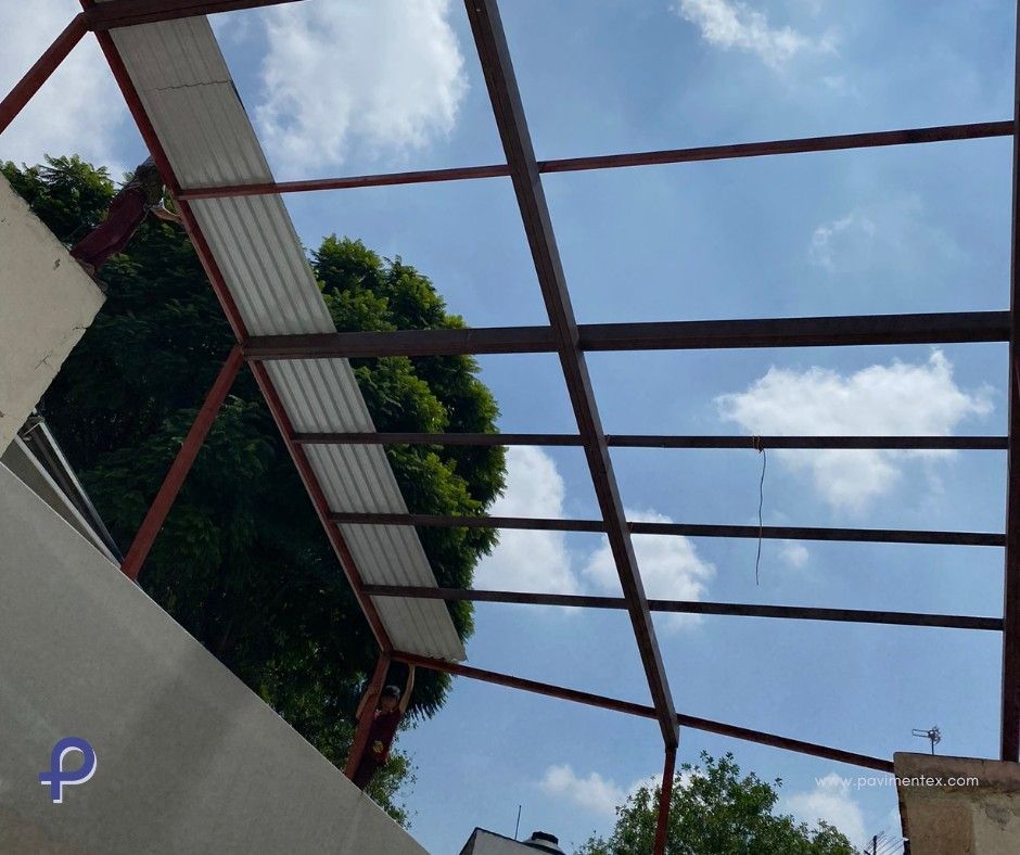 Estructura de techo con marco de metal contra un cielo azul nublado, con paneles corrugados blancos y vegetación circundante.