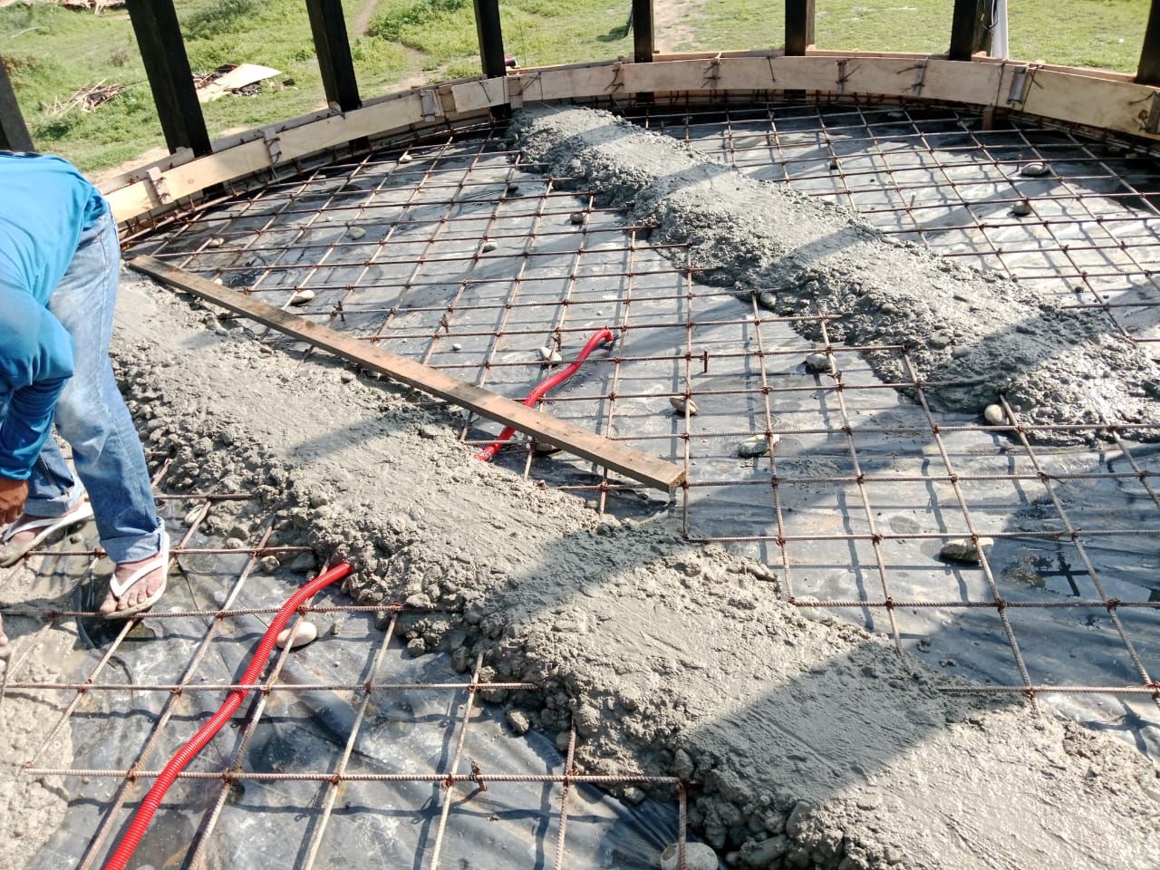 Construcción de un piso circular de concreto; rejilla de varillas de refuerzo, concreto fresco y trabajador con camisa azul.