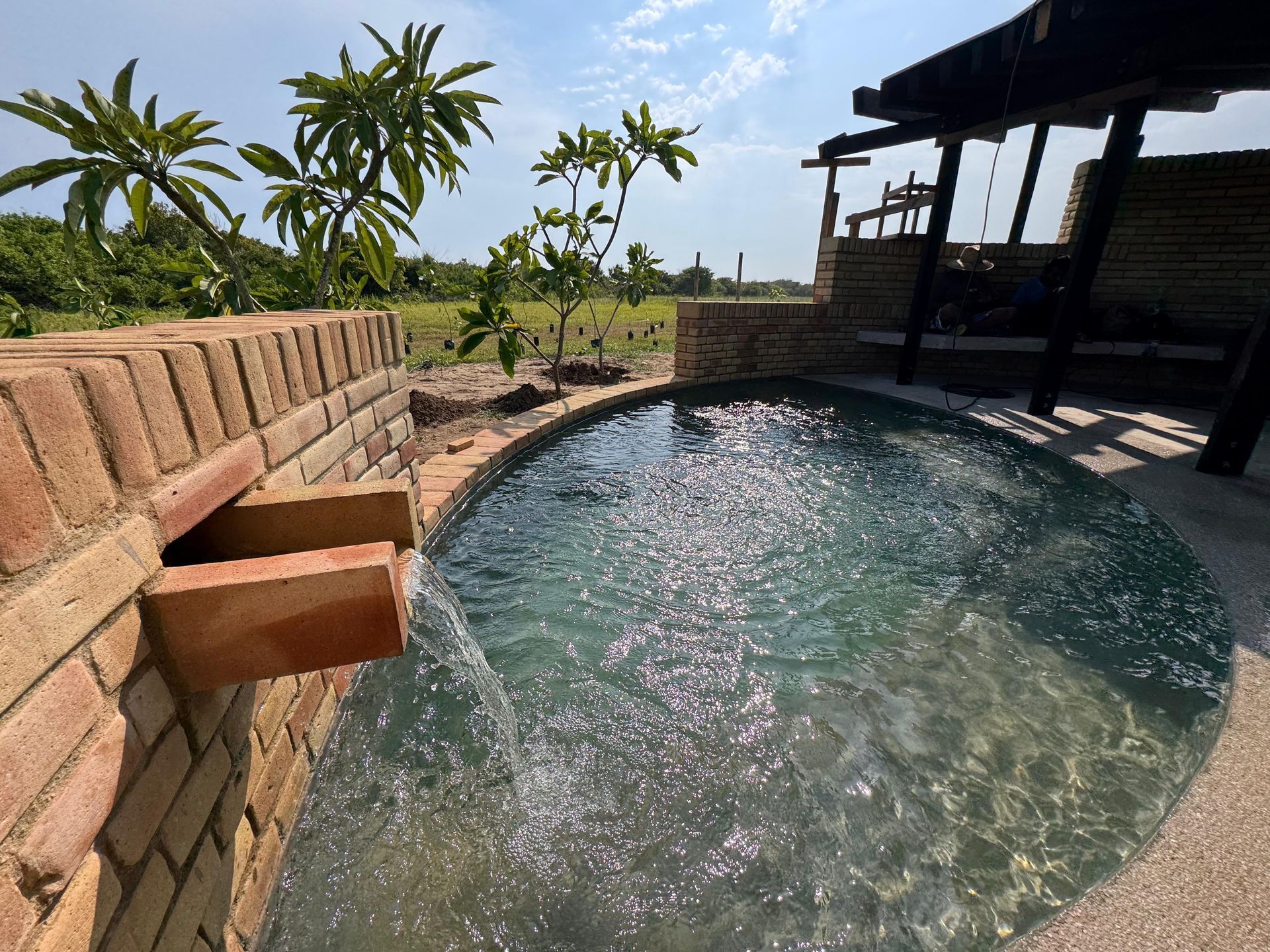 Piscina al aire libre revestida de ladrillo con cascada de agua; ubicada en un entorno tropical soleado.