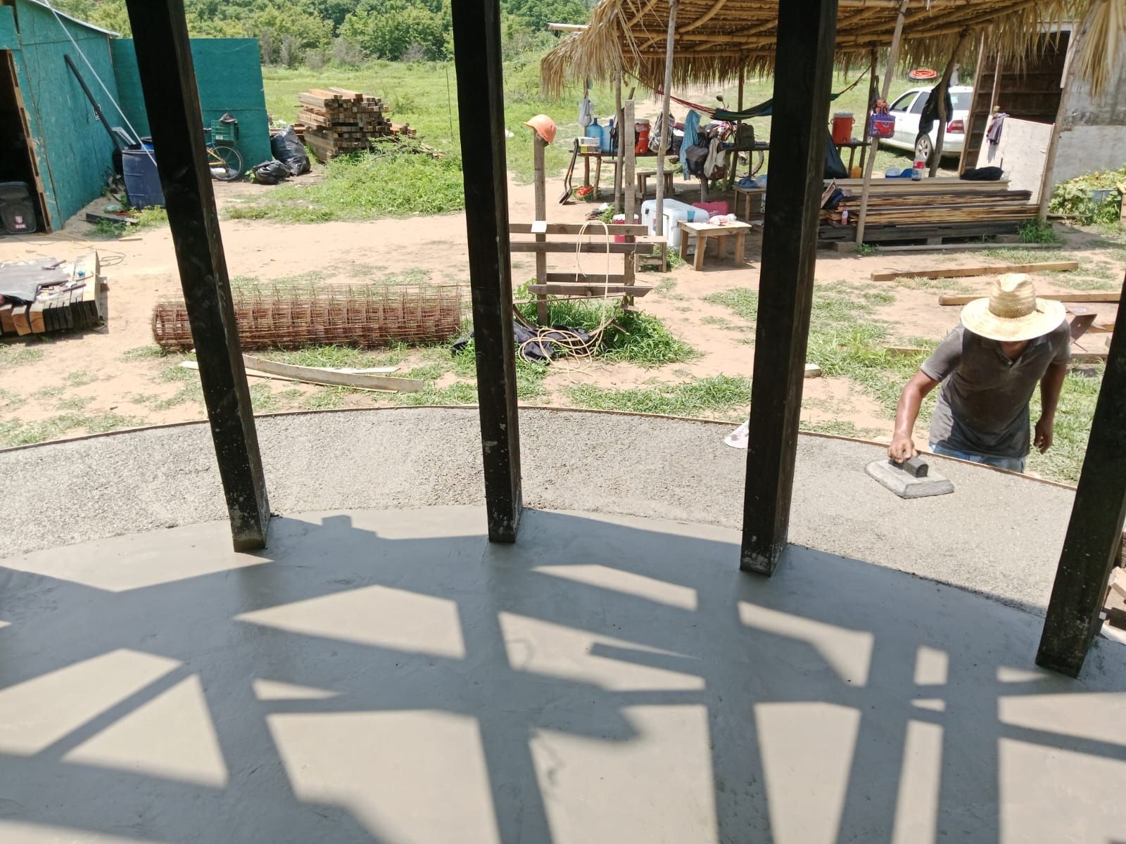 Persona alisando piso de concreto alrededor de postes de madera al aire libre; día soleado.