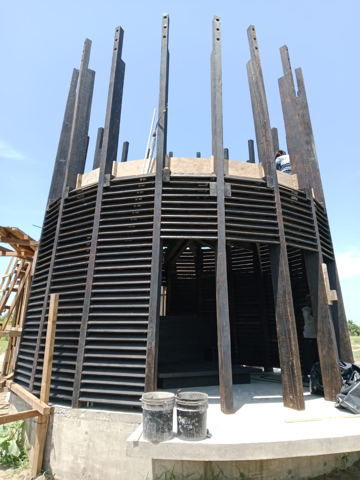 Construcción de una estructura de madera oscura, con vigas verticales y listones horizontales. Exterior con cielo azul.