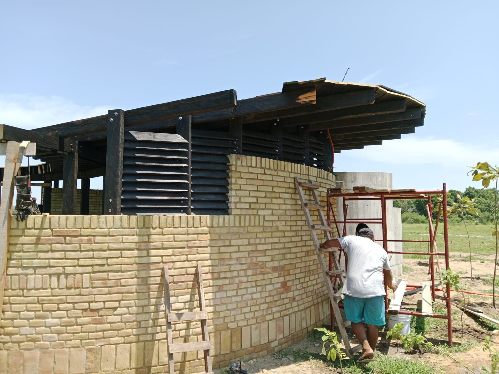 Hombre subiendo escalera junto a edificio de ladrillo en construcción; vigas de madera y listones negros, cielo azul.