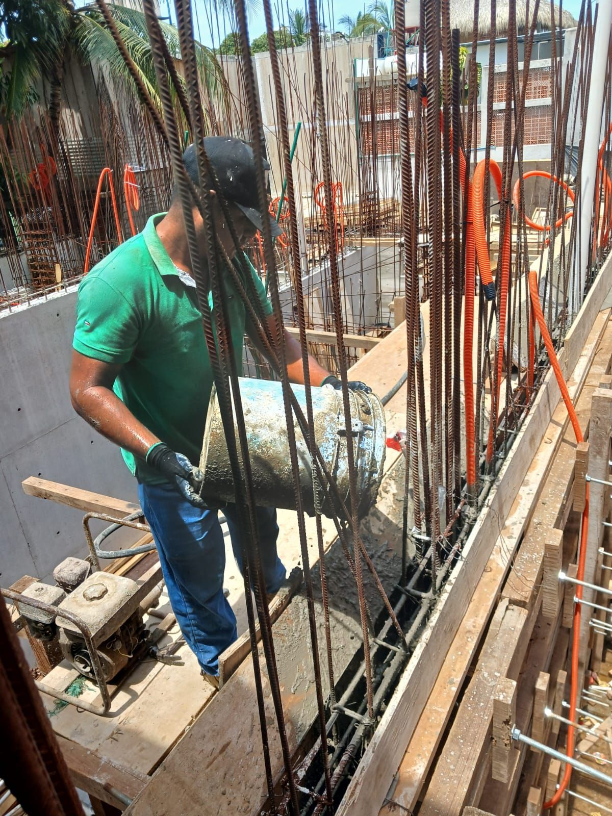 Obrero de la construcción vierte cemento en un encofrado, rodeado de barras de refuerzo y líneas de servicios públicos.