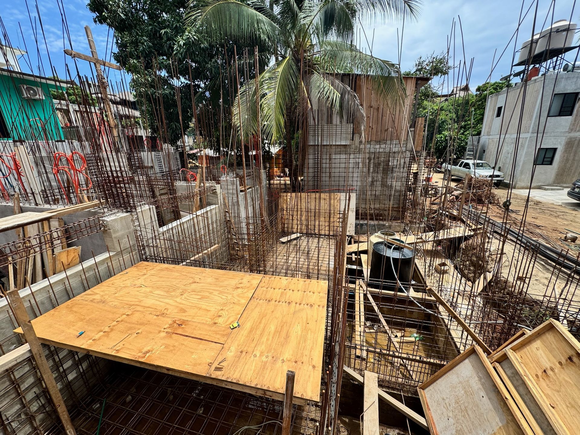 Sitio de construcción con encofrados de hormigón, varillas de refuerzo expuestas, paneles de madera y estructura parcialmente construida.