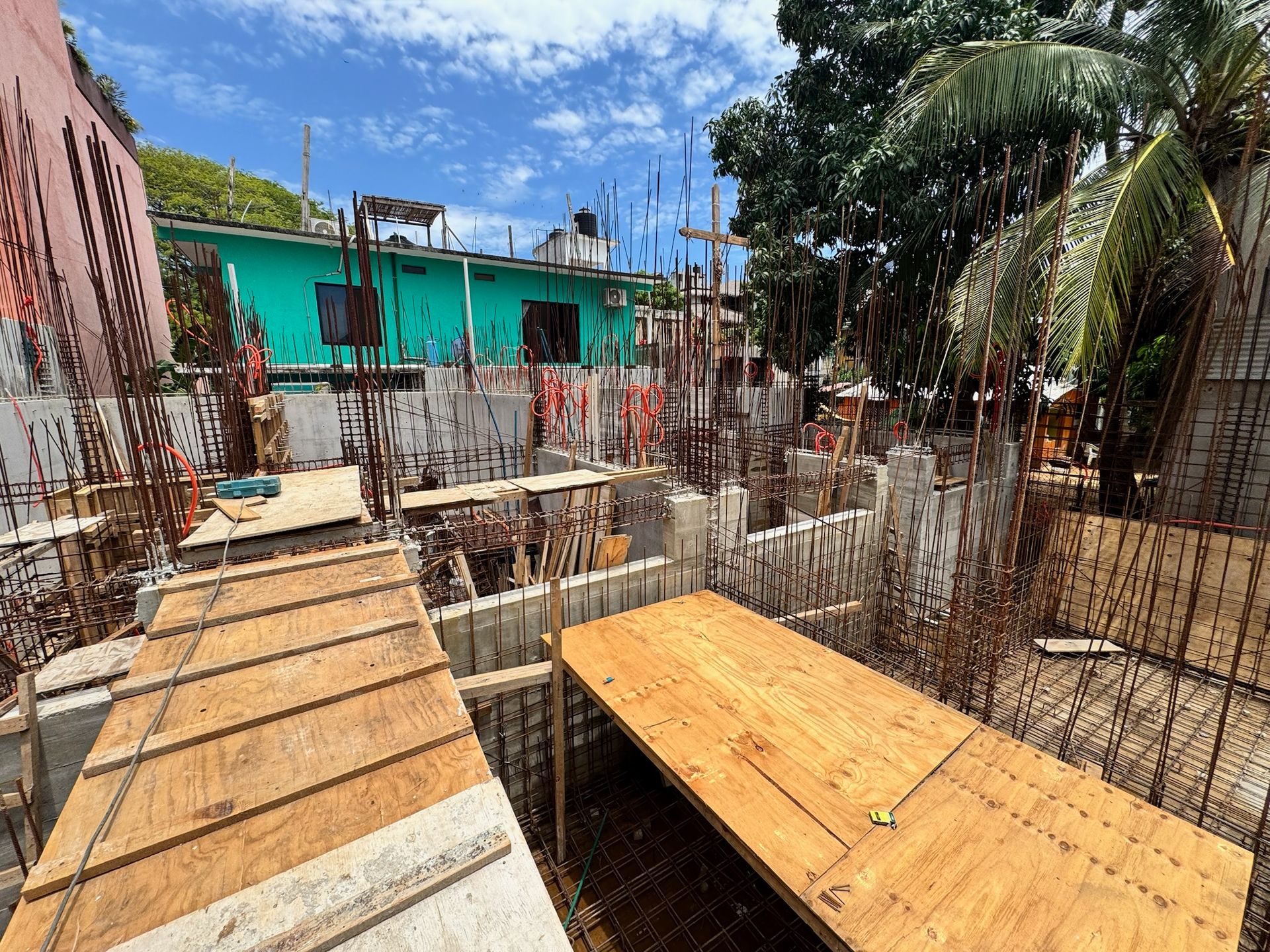 Sitio de construcción con plataformas de madera, paredes de hormigón, barras de refuerzo y un edificio verde azulado al fondo bajo un cielo azul.