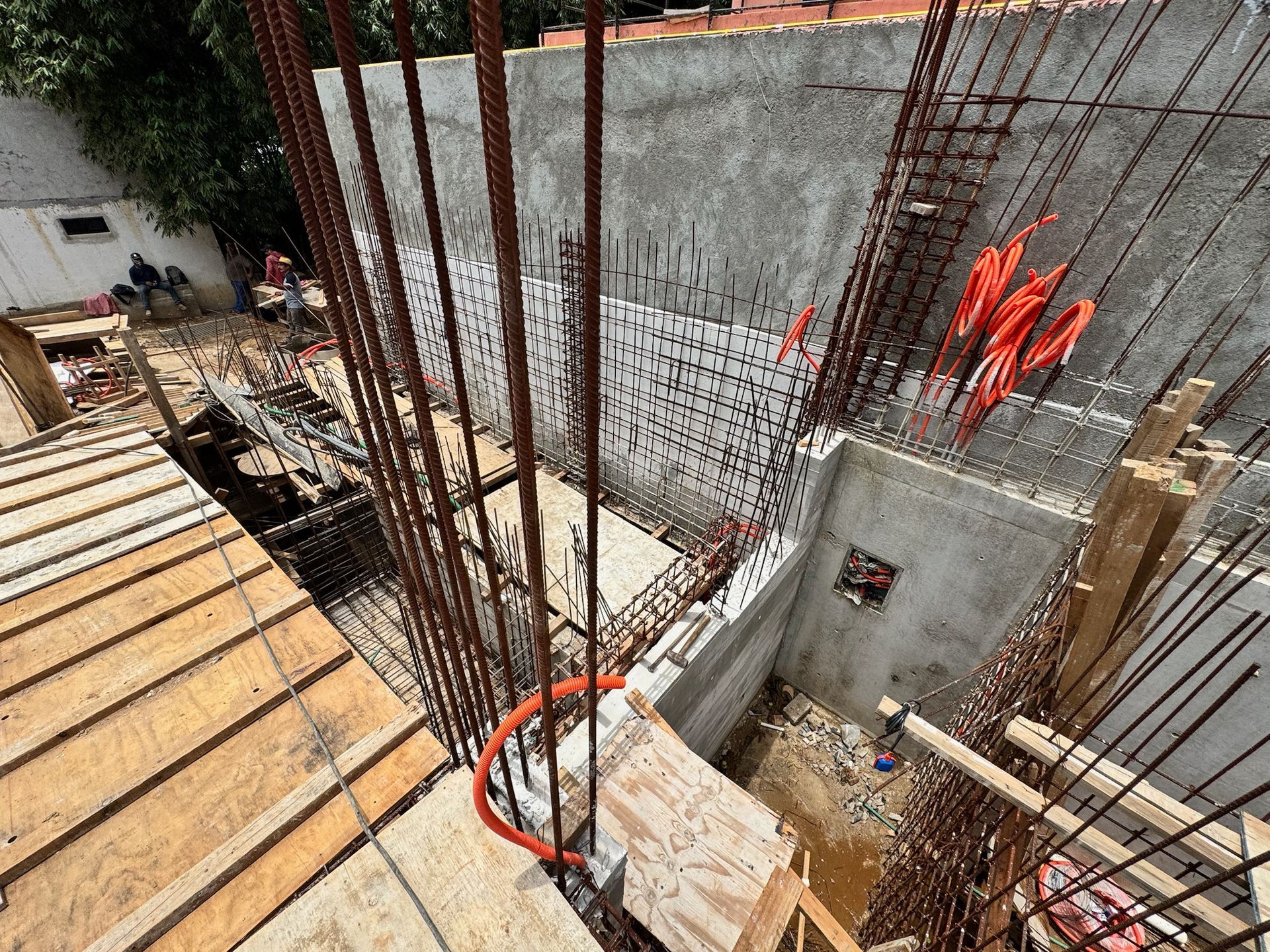 Obra: muros de hormigón, estructura de varillas de refuerzo, encofrados de madera, conductos eléctricos de color naranja y cielo nublado.