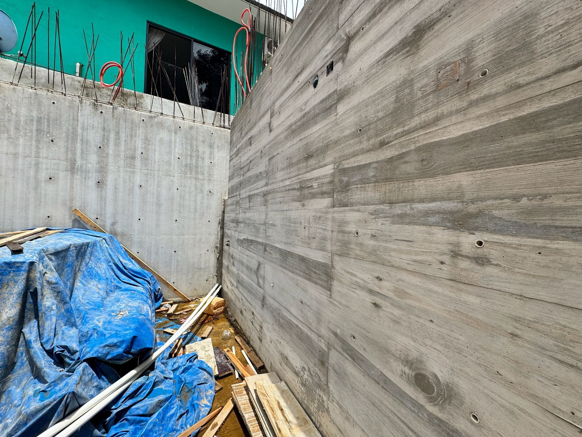 Muros de hormigón en construcción, con un edificio turquesa al fondo. Escombros y una lona azul en la base.