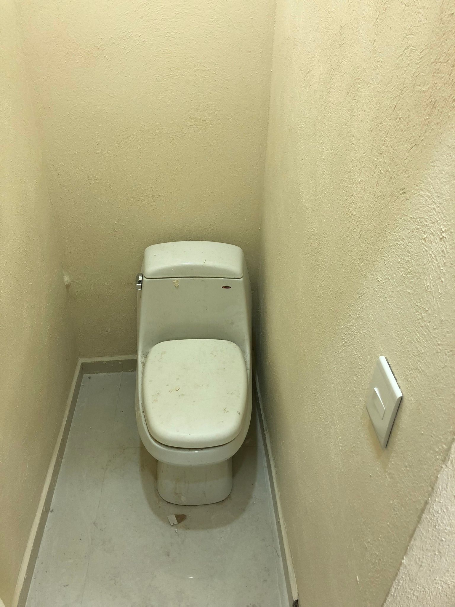 Inodoro en un baño pequeño con paredes beige. Suelo gris claro, inodoro blanco con la tapa bajada.