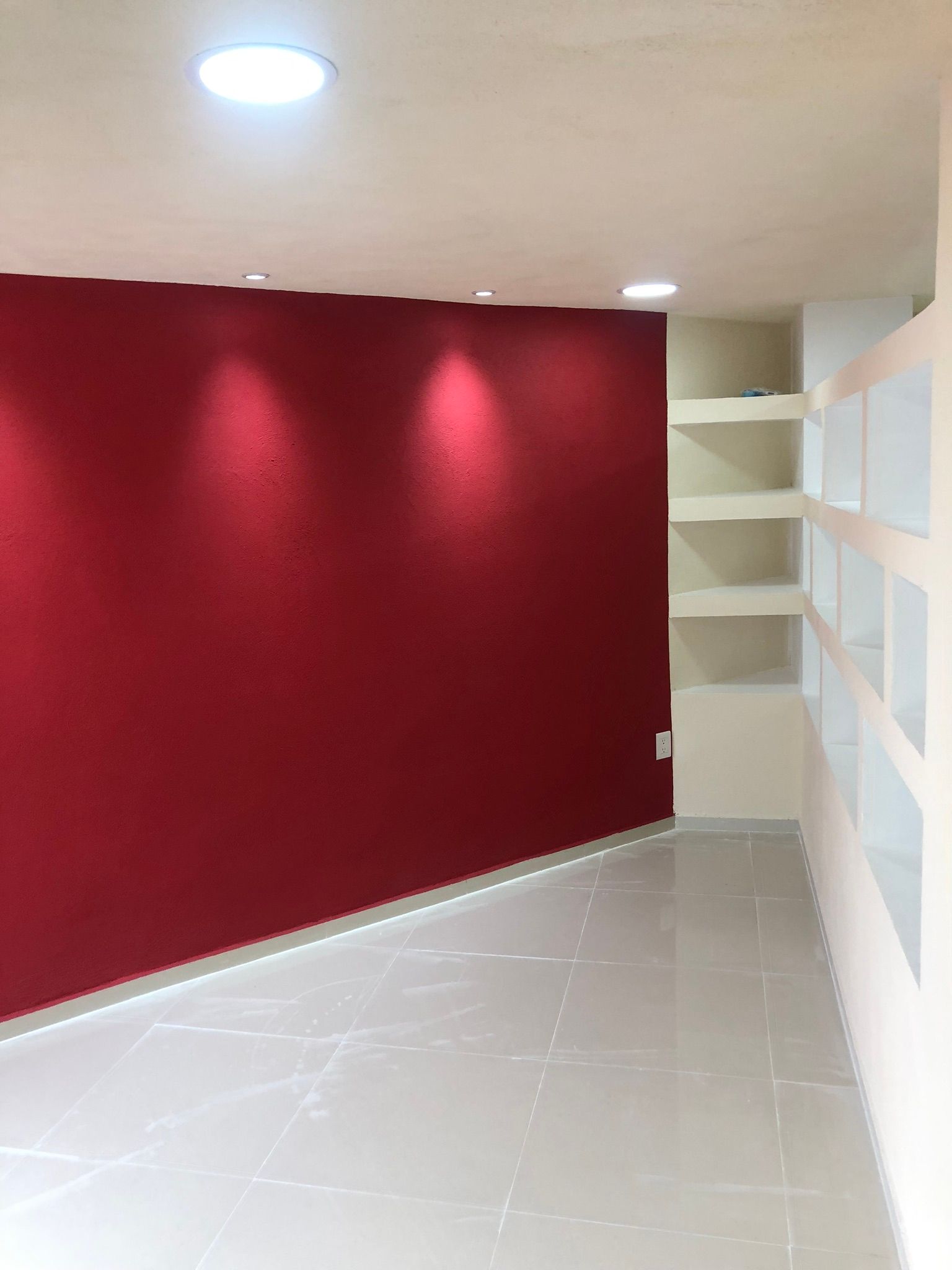 Habitación vacía con pared de acento rojo, estantes empotrados y piso de baldosas blancas.