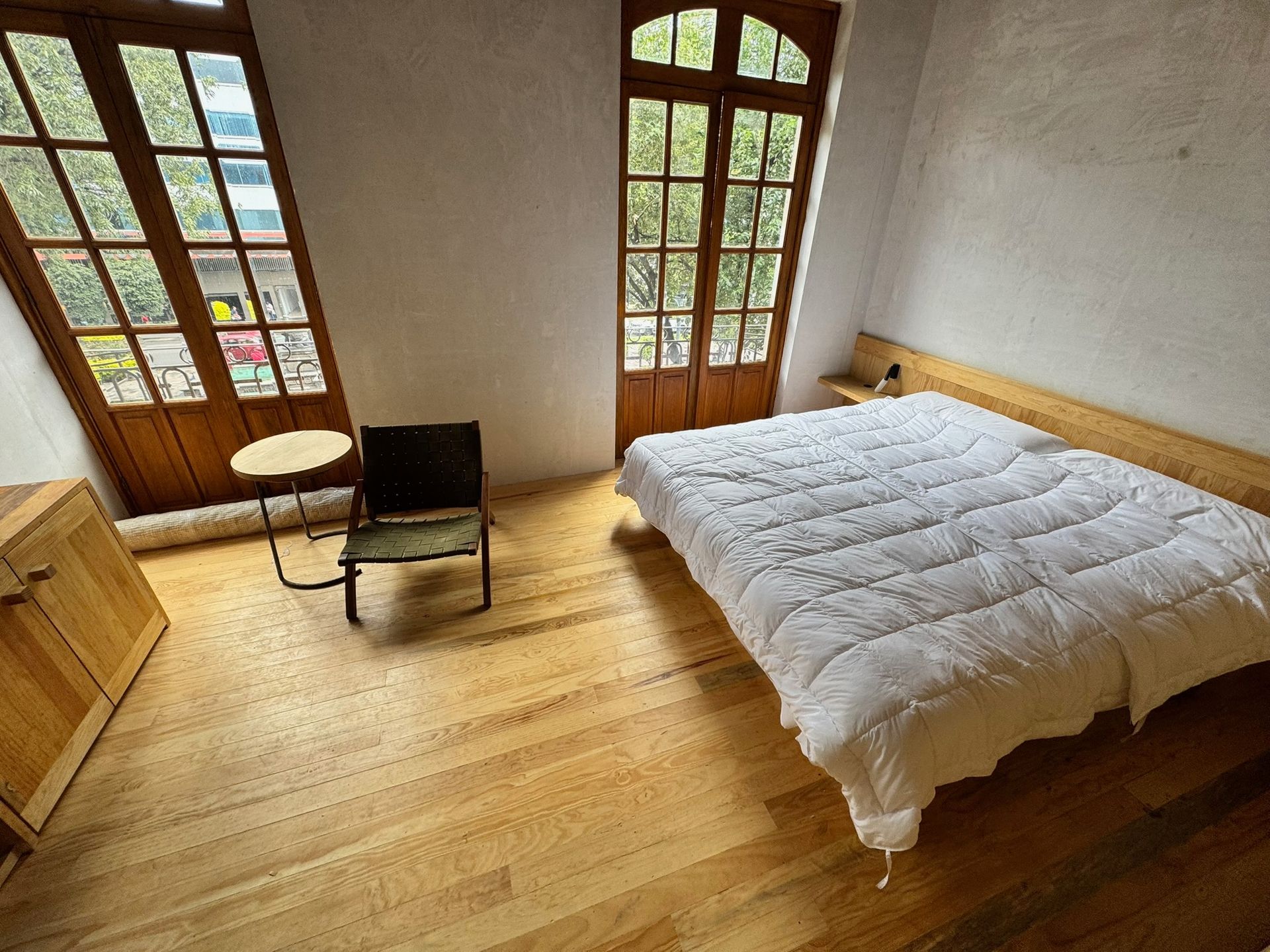 Dormitorio con suelo de madera, ventanas y cama con edredón blanco. Incluye silla y mesa pequeña.