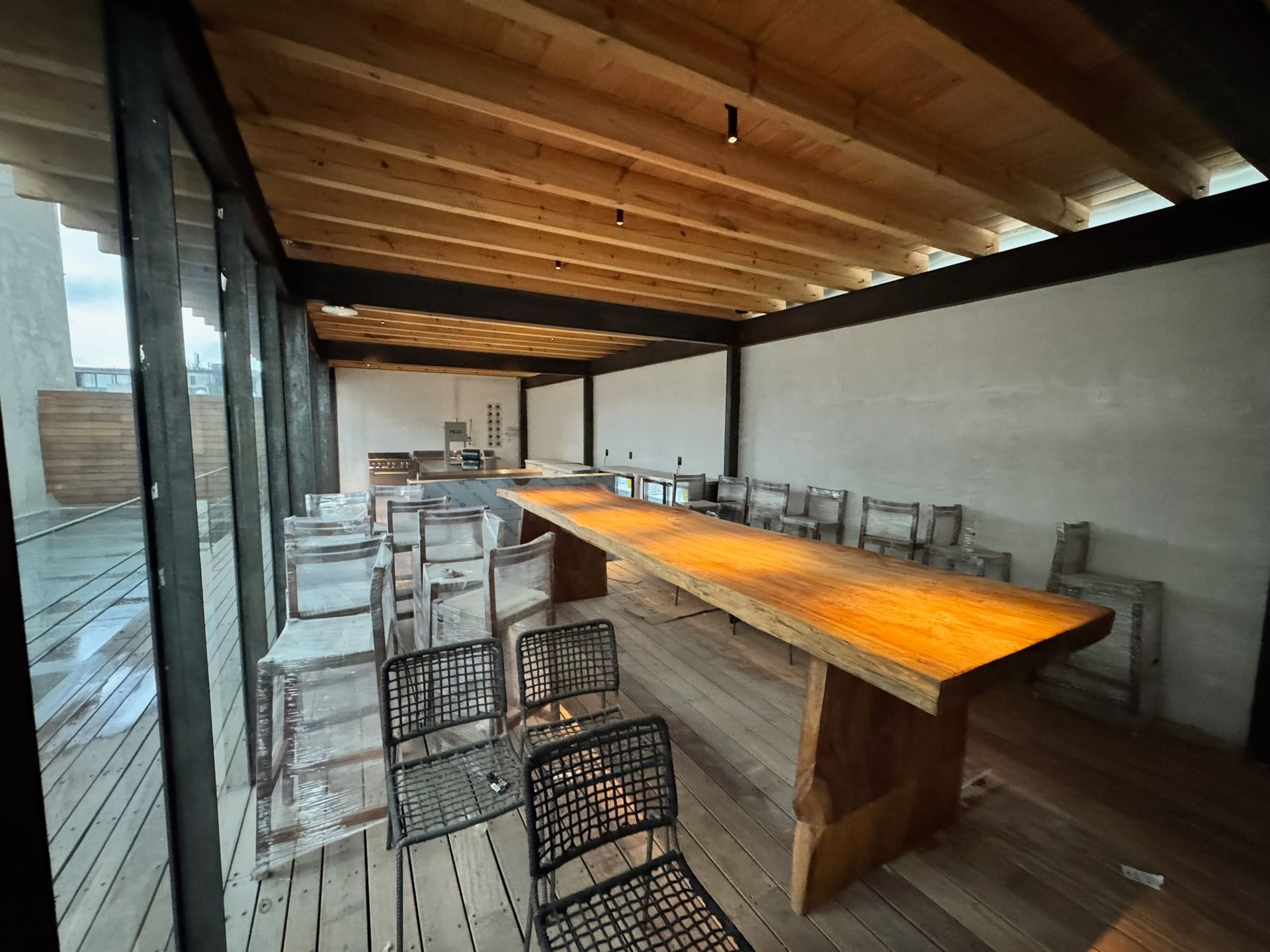 Mesa larga de madera con sillas en una habitación al aire libre, iluminada por el sol, con paredes de cristal y techo de madera.
