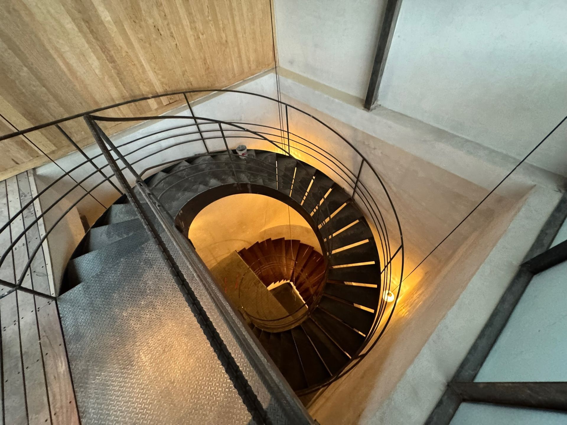 Escalera de caracol con peldaños y barandilla metálica, que asciende hacia una fuente de luz.