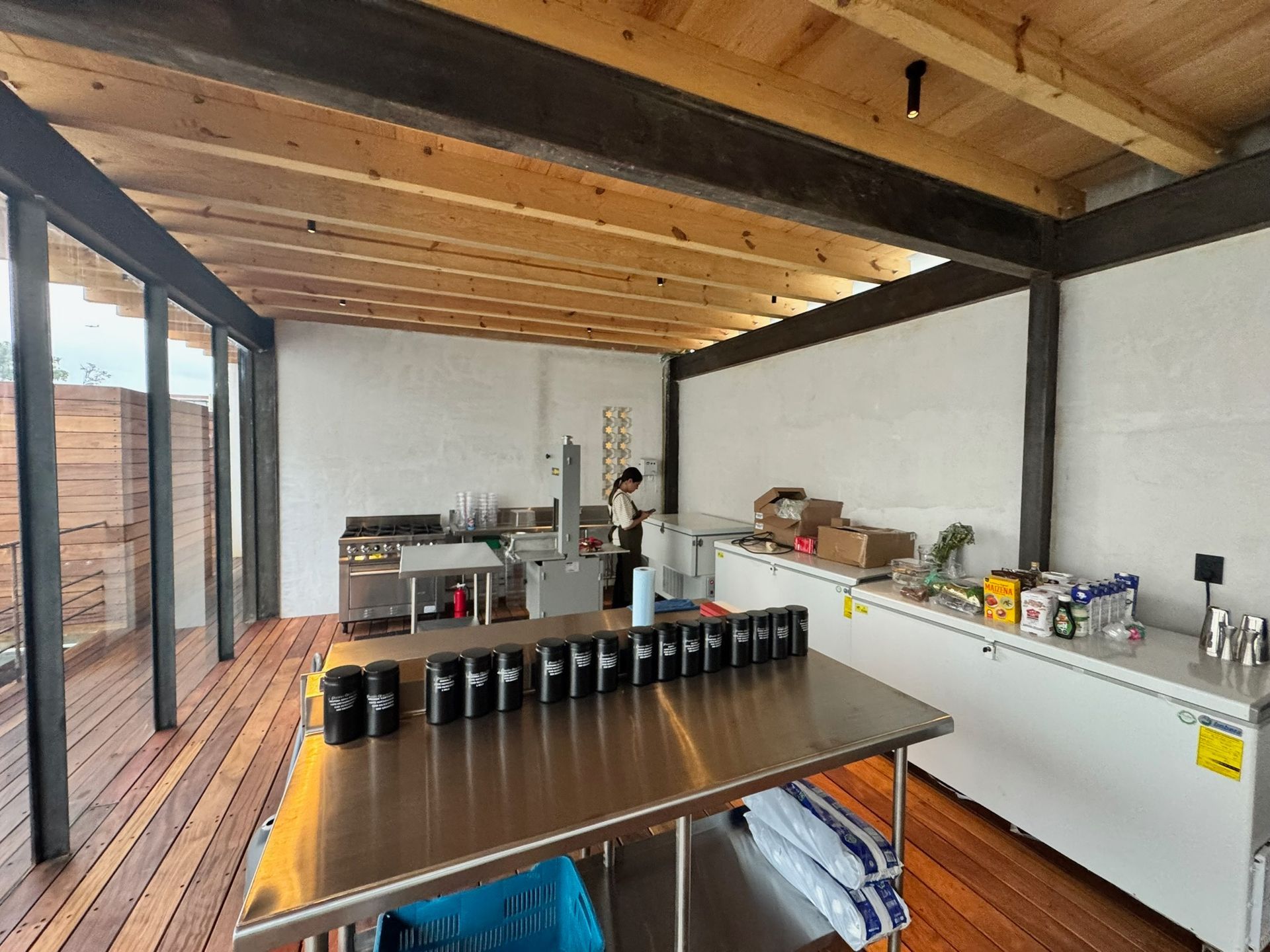 Una cocina comercial con mesas de acero inoxidable, una persona trabajando y una terraza con tablones de madera.