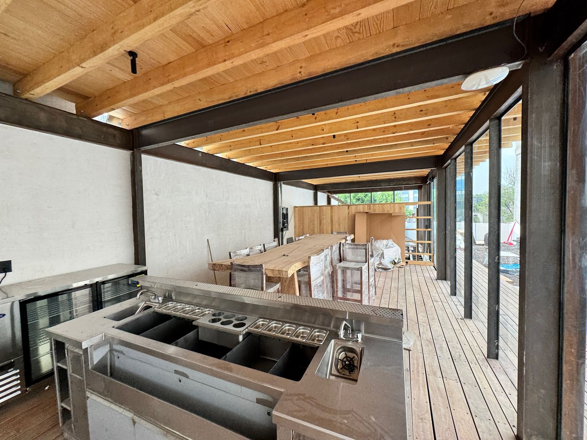 Bar exterior en construcción: mostradores de acero inoxidable, vigas de madera, paredes de cristal.