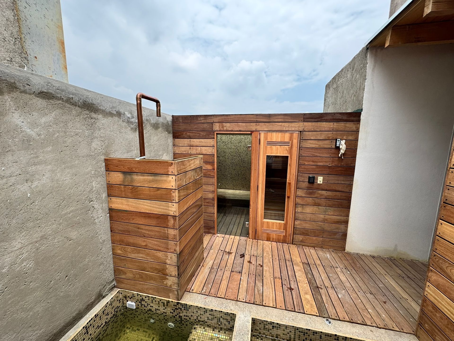 Ducha exterior de madera y sauna en un patio, con un cielo gris encima.