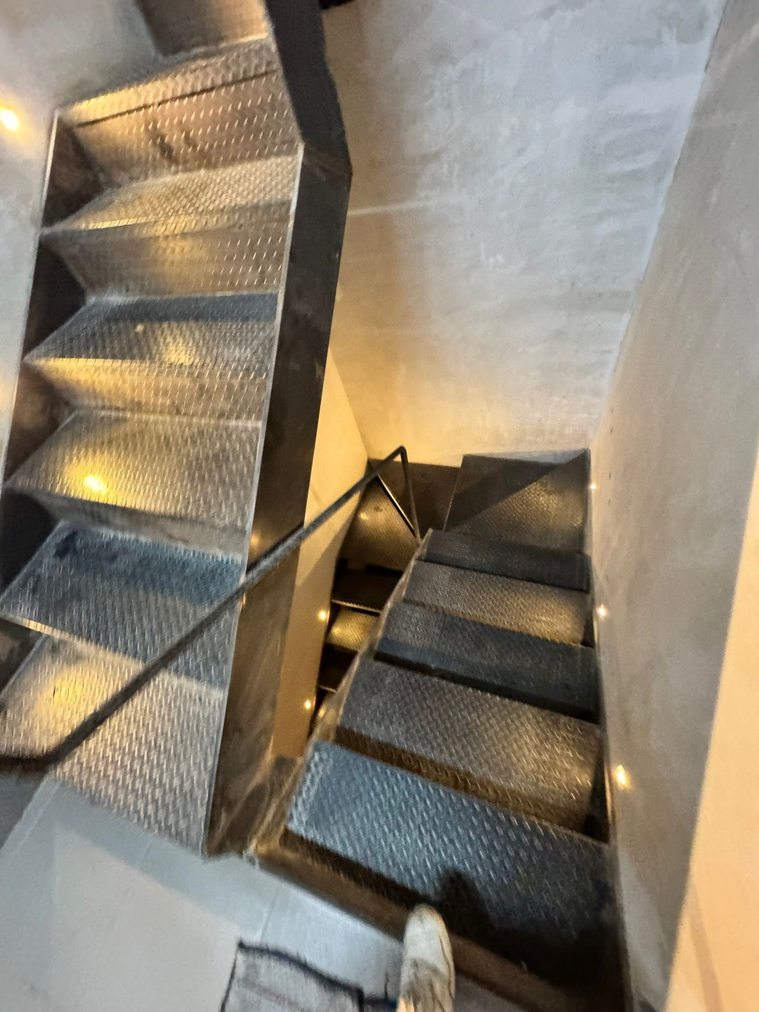 Escalera metálica con escalones texturizados, que desciende de arriba a la derecha hacia abajo a la izquierda. Iluminación tenue.