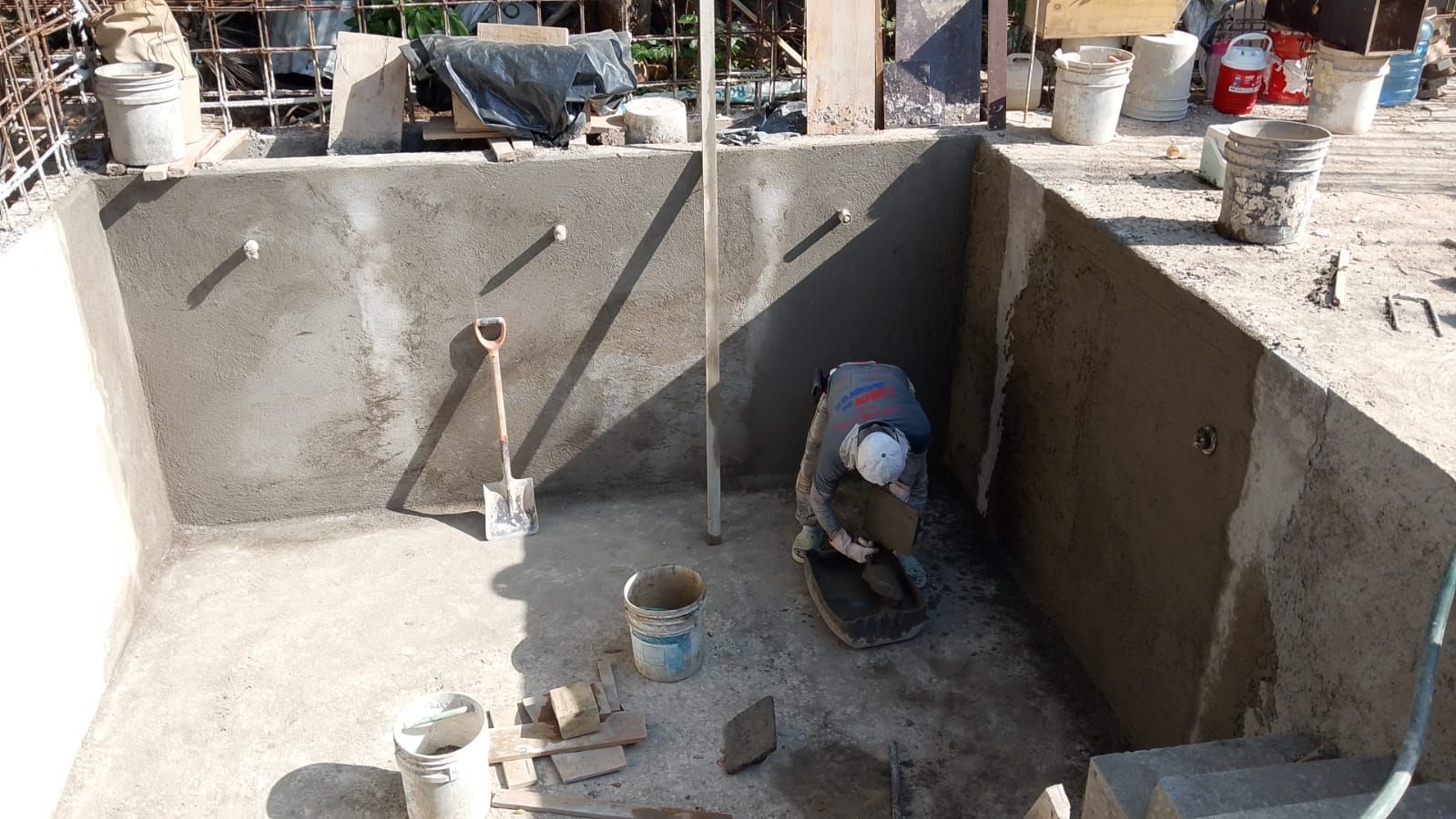 Un obrero enyesando el interior de una piscina de hormigón. Cubos, herramientas y materiales de construcción los rodean.