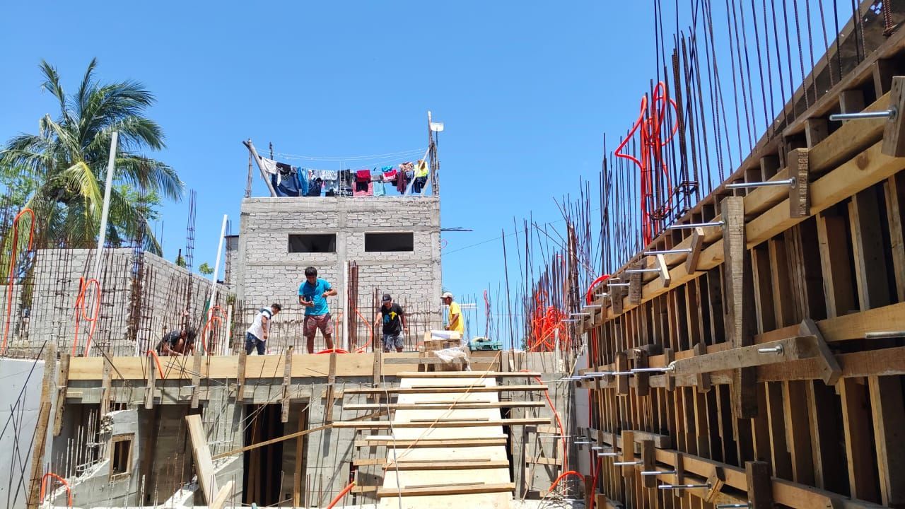 Obra en construcción con obreros, encofrados de madera, hormigón y un edificio con ropa tendida.