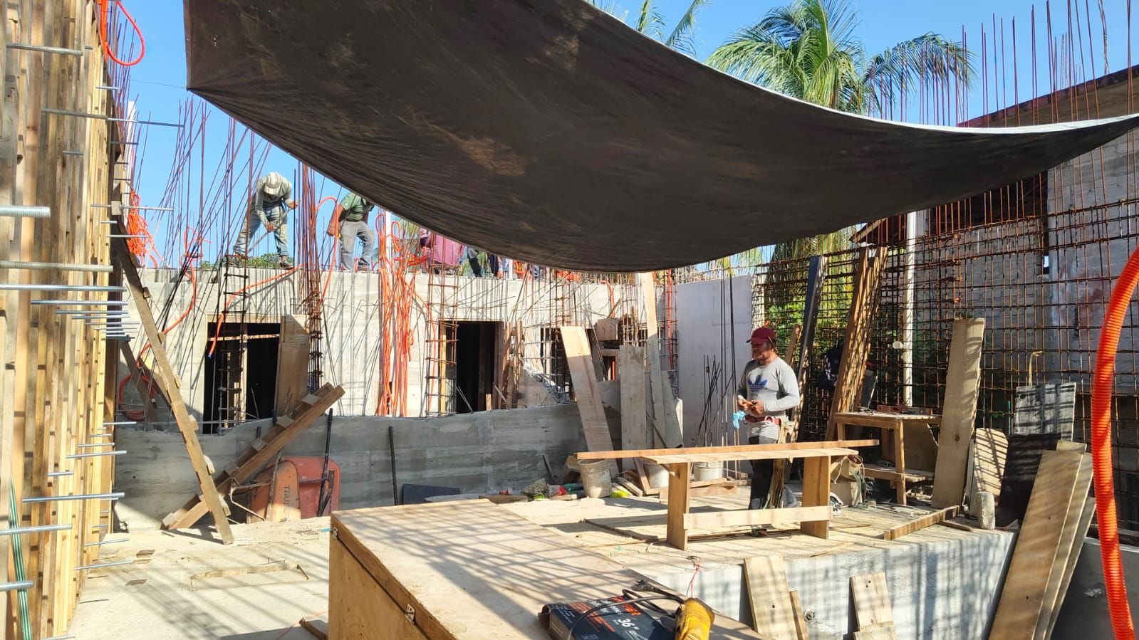 Sitio de construcción con trabajadores, encofrados de hormigón y toldo parasol.