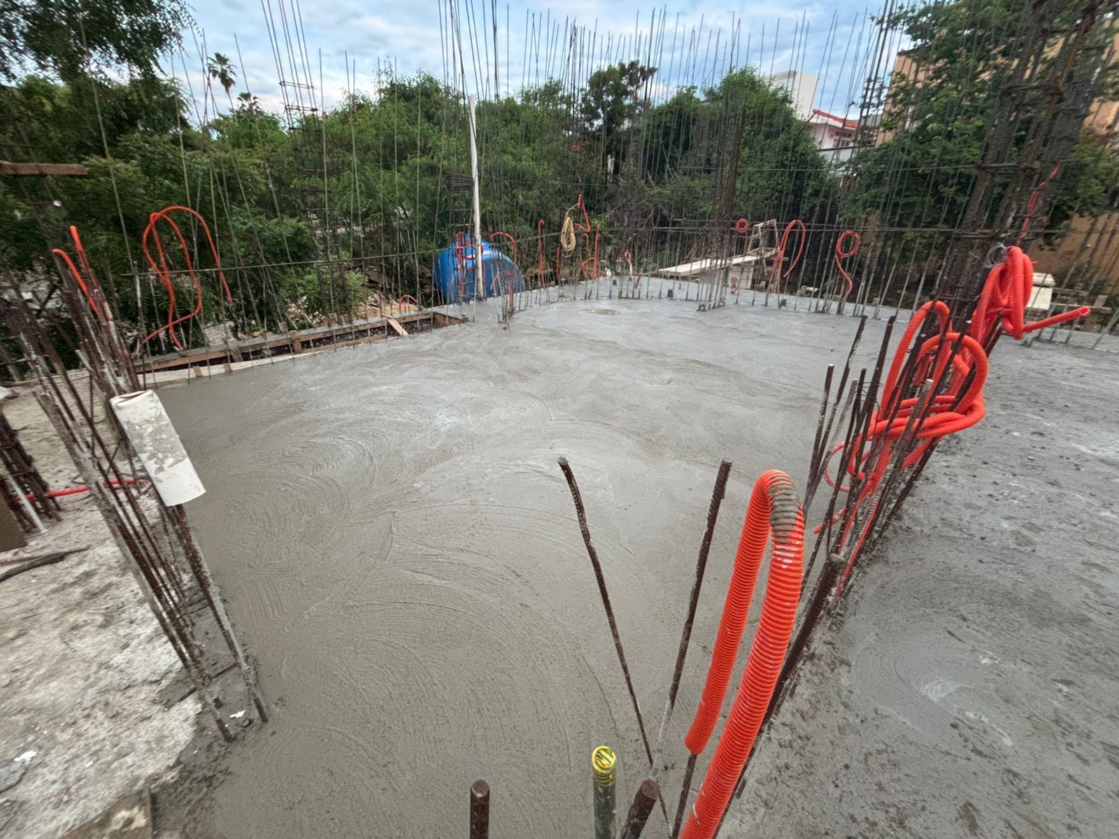 Cimentación de hormigón recién vertida en una obra en construcción, con barras de refuerzo expuestas y conductos de color naranja.