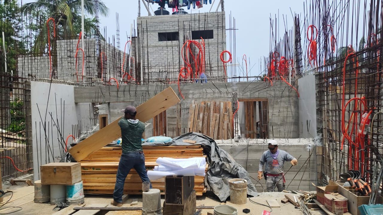 Obreros de la construcción transportan madera en una obra con estructura de hormigón a la vista y tuberías rojas.