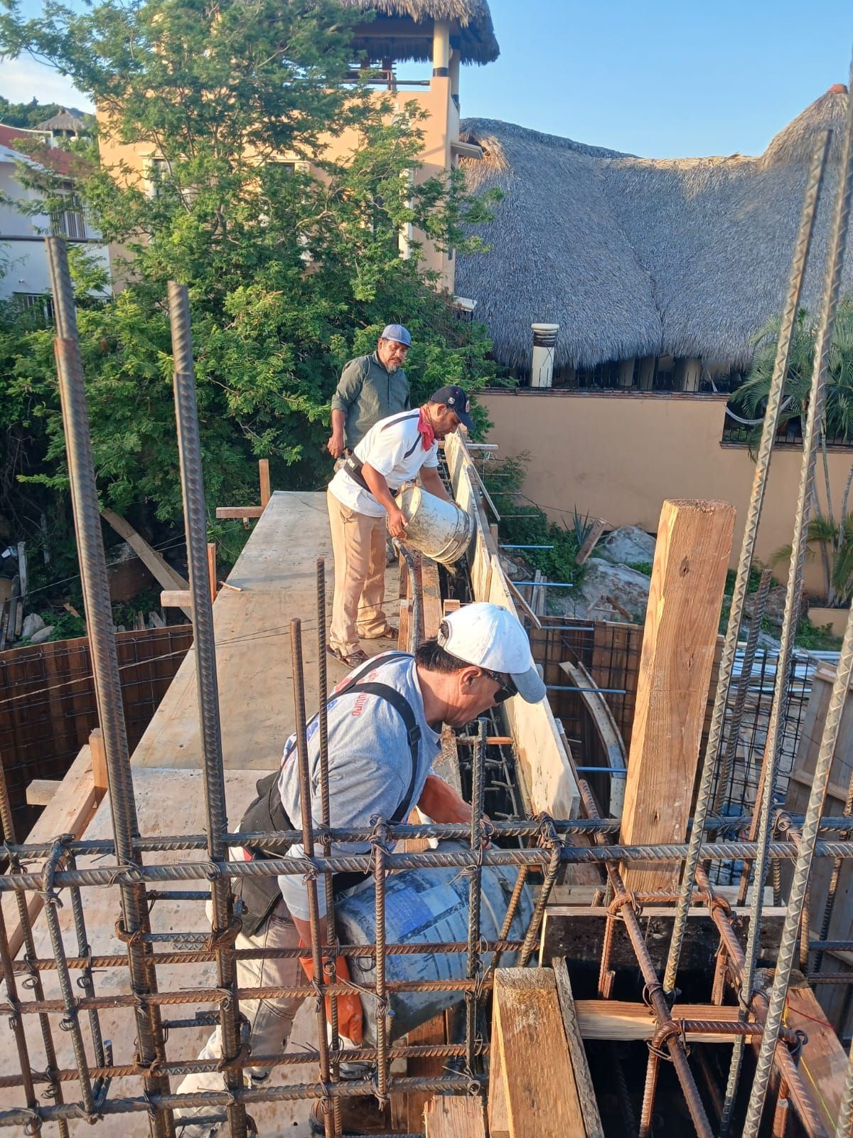 Obreros de la construcción vertiendo hormigón; entorno al aire libre, luz solar, barras de acero, pantalones color canela, camisas blancas y grises.