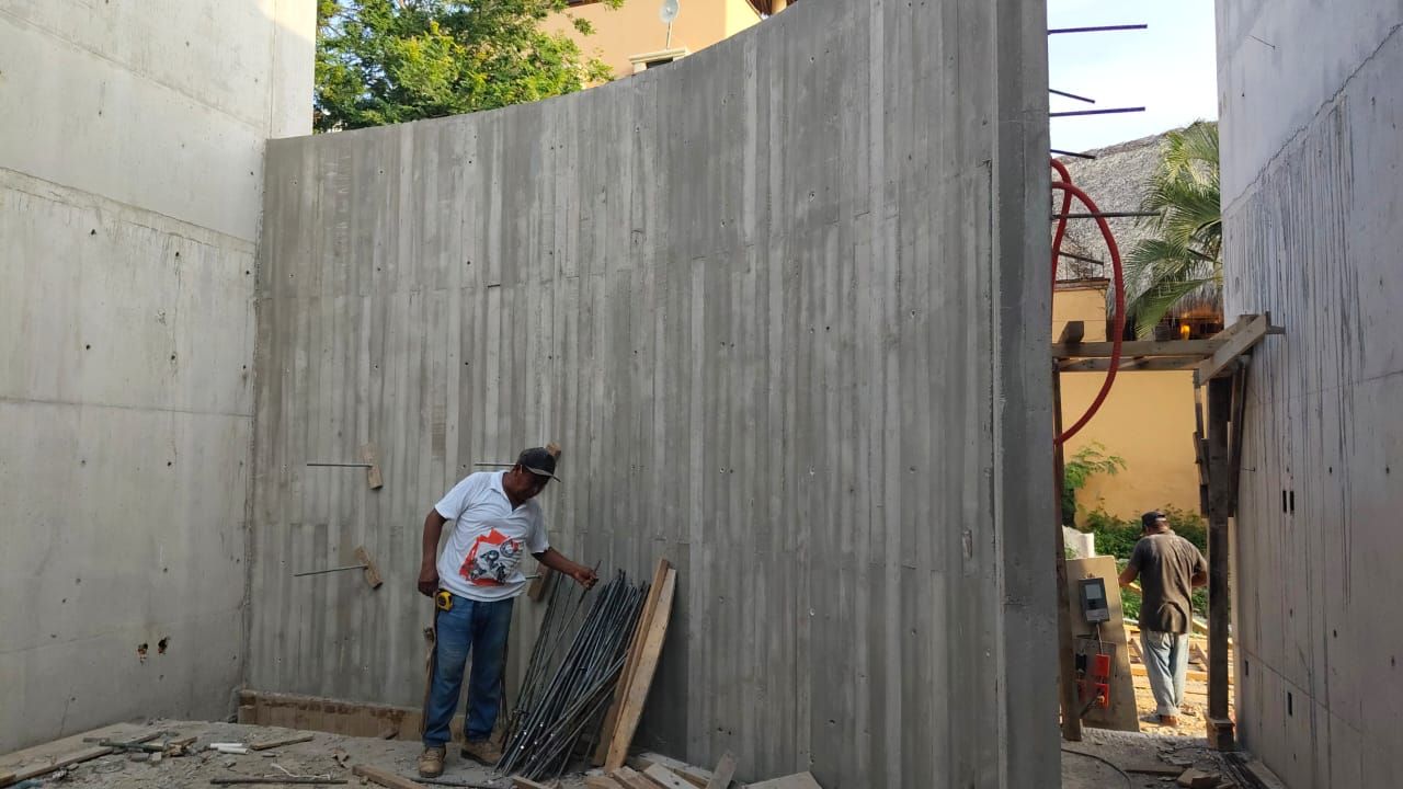 Un obrero inspecciona un muro de hormigón curvo. Al fondo se ven varillas de refuerzo y otros trabajadores.
