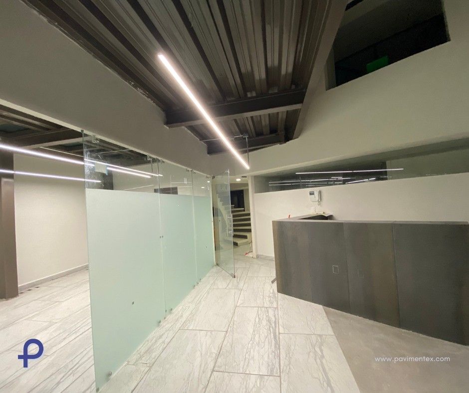 Interior de oficina moderno con paredes de vidrio, mostrador de recepción y escaleras al fondo.