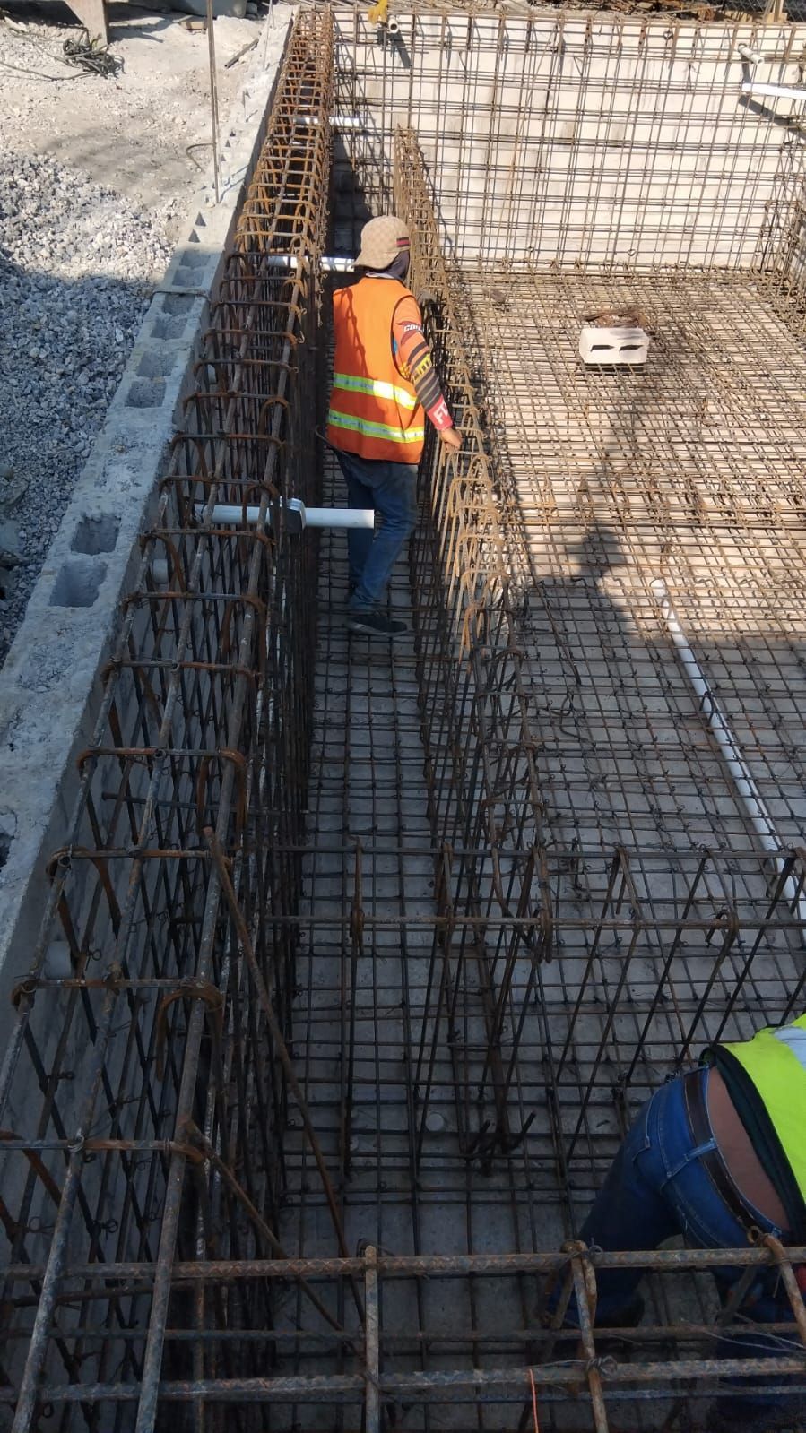Obreros de la construcción en un encofrado de hormigón, trabajando con varillas de refuerzo. Una persona lleva un chaleco naranja.