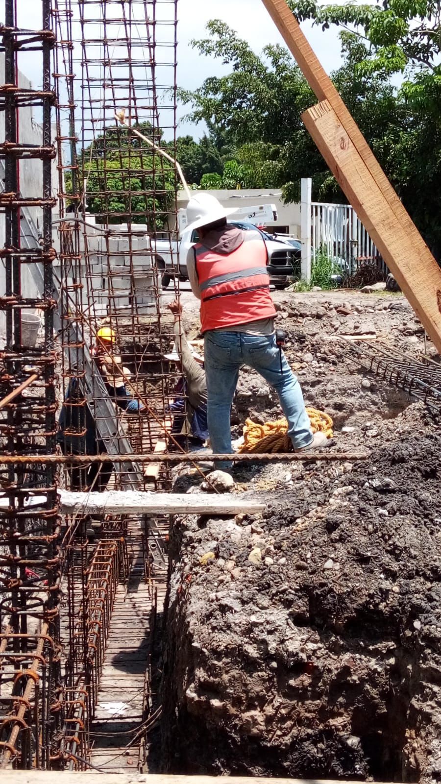 Obrero con chaleco naranja, de pie en una zanja, inspeccionando una estructura de varillas de refuerzo. Al aire libre, a plena luz del día.