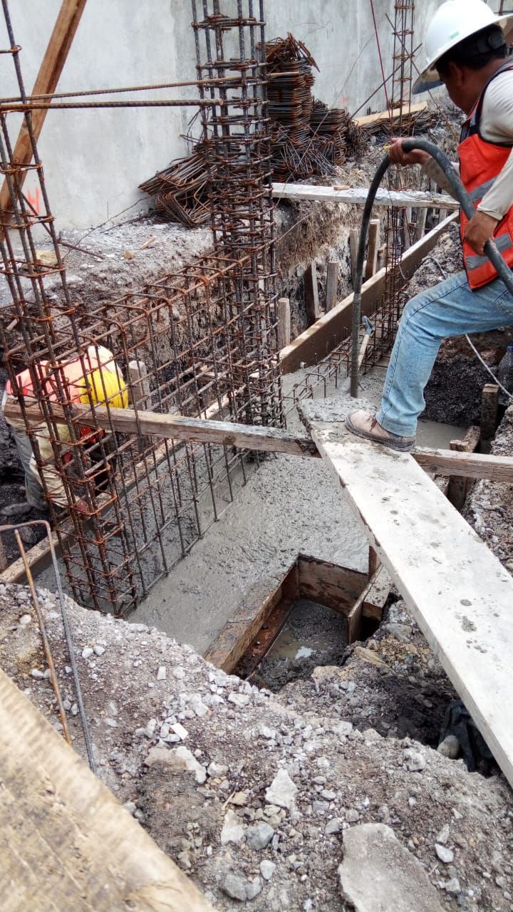 Obreros vierten hormigón en una cimentación. Un trabajador usa una manguera. Se ven varillas de refuerzo y un encofrado de hormigón.