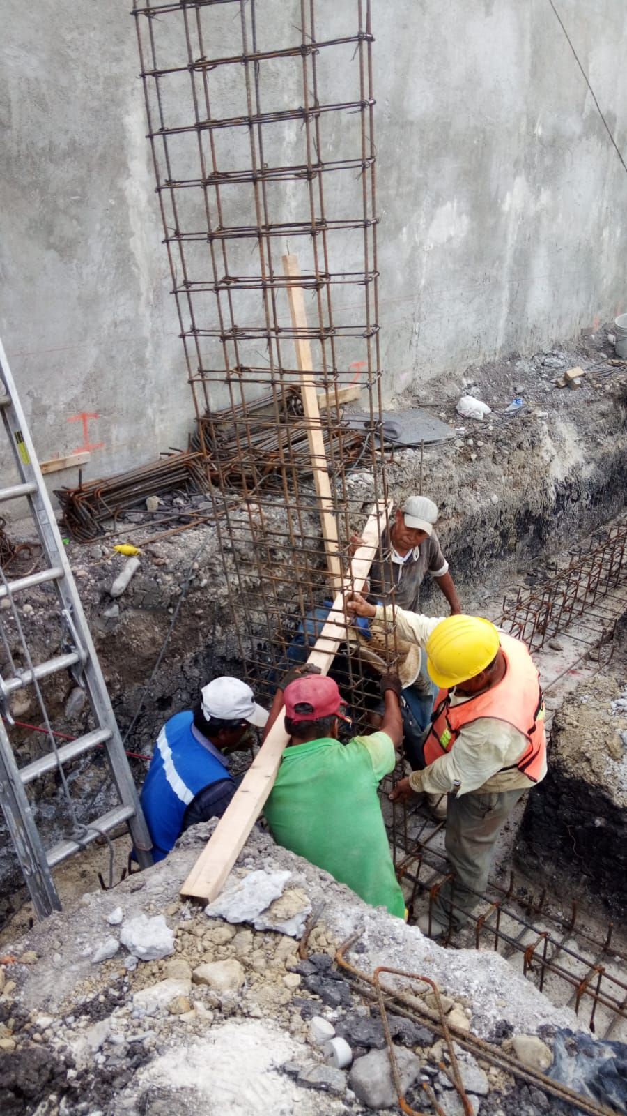Obreros instalan varillas de refuerzo para una columna de hormigón en una zanja. Los tonos tierra dominan la escena.