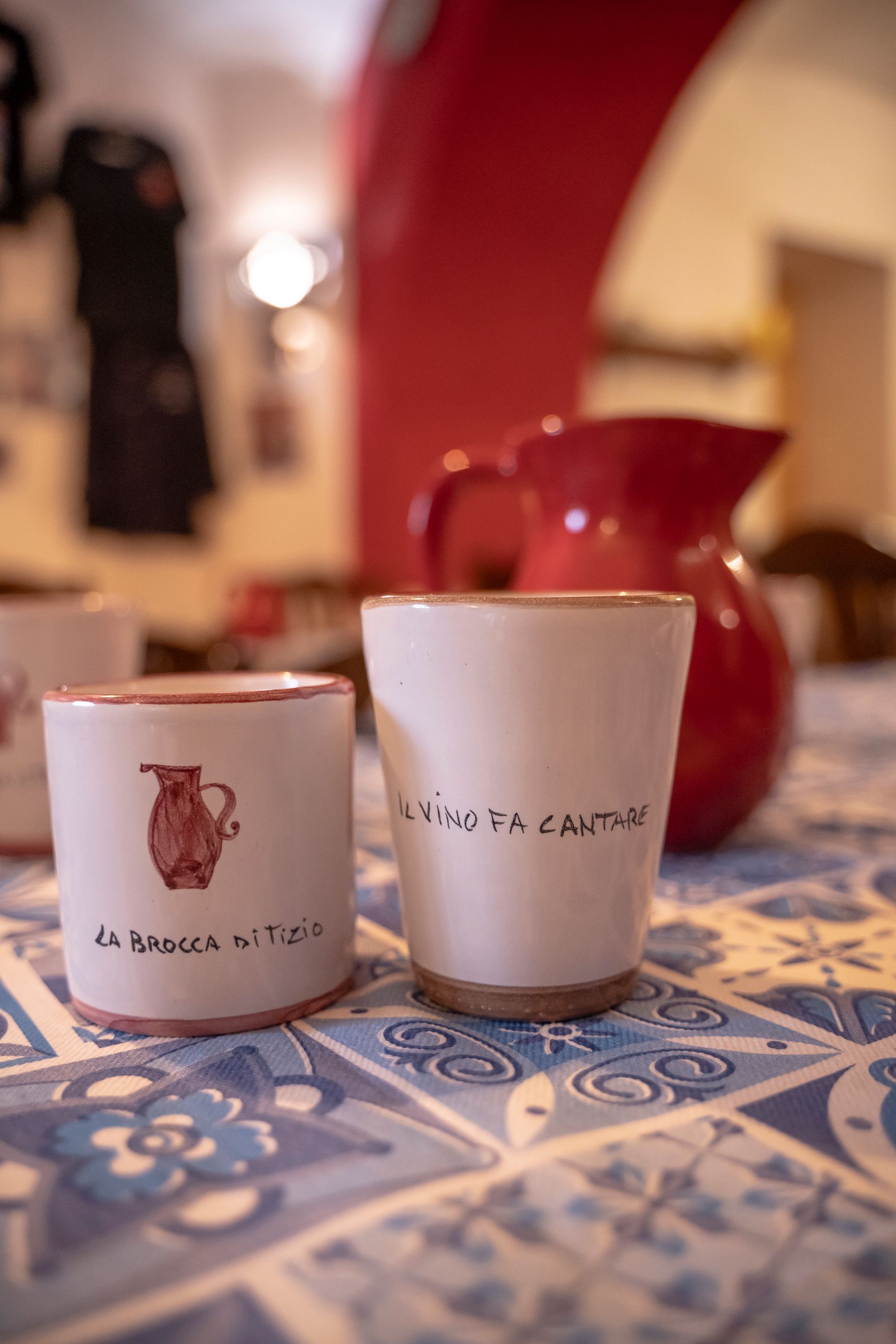 Due tazze di ceramica con testo scritto a mano su una tovaglia a motivi blu e bianchi, con una brocca rossa sullo sfondo.