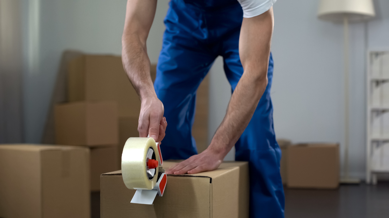 Person in blue jumpsuit taping a cardboard box. Boxes stacked in the background.
