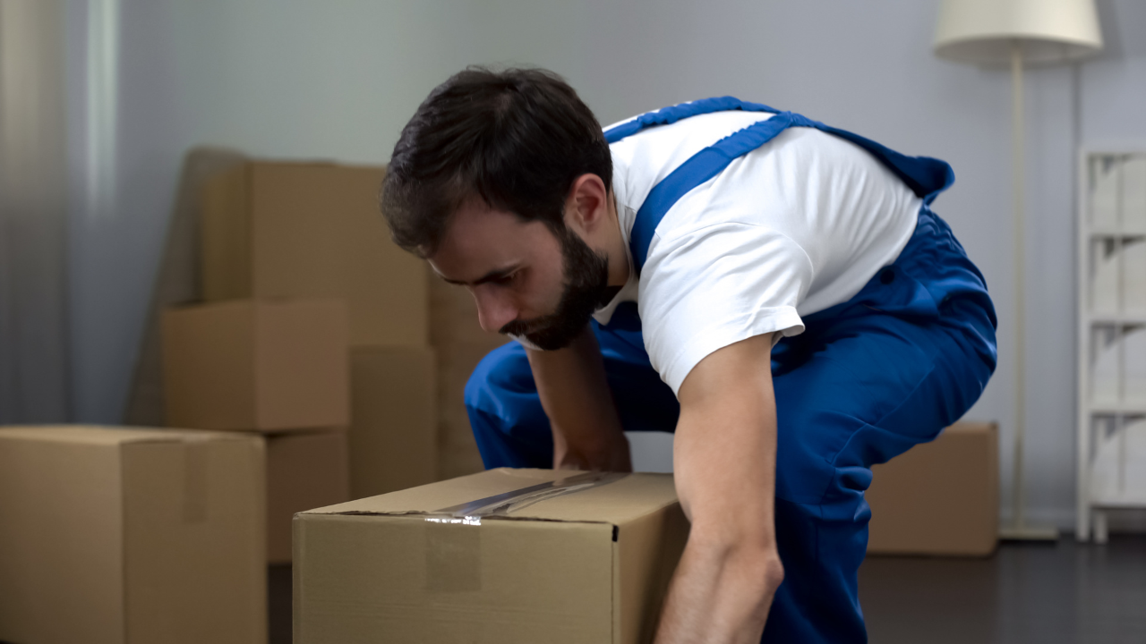 Man in blue overalls lifting a cardboard box, indoors, boxes stacked in the background.