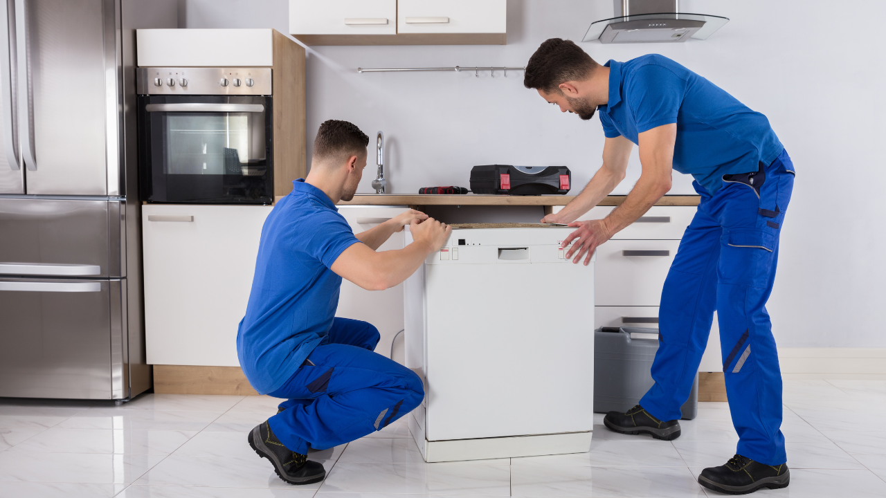 Two technicians in blue uniforms install a dishwasher in a kitchen.