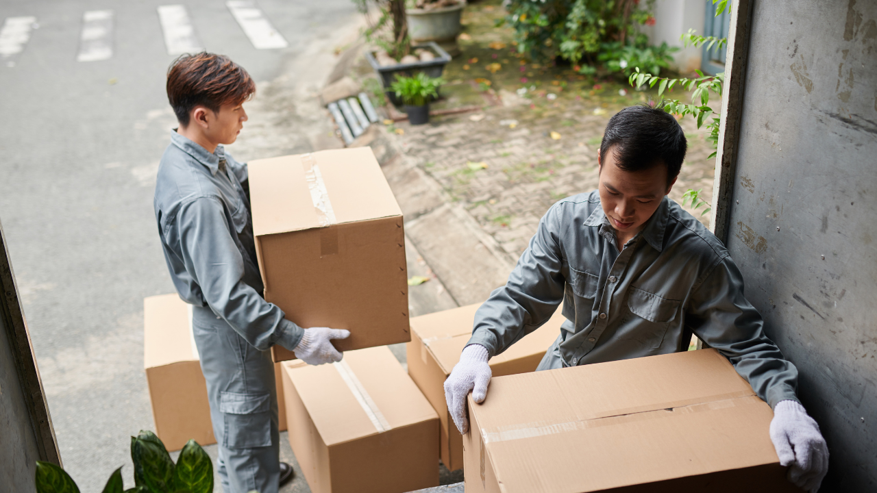 Two people in work clothes carry moving boxes into a doorway.